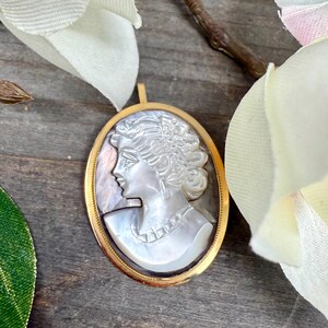 May include: A gold-toned oval pendant with a cameo of a woman's profile carved from mother-of-pearl. The cameo is set in a gold-toned frame.