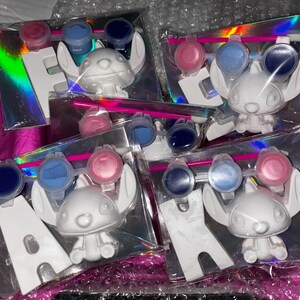 May include: Five paint-your-own ceramic figurines of Stitch from Lilo & Stitch. Each figurine comes with three small paint pots, a paintbrush, and a letter. The letters spell out the word "HAWAII".