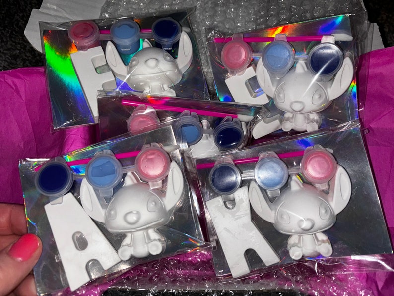 May include: Five paint-your-own ceramic figurines of Stitch from Lilo & Stitch. Each figurine comes with three small paint pots, a paintbrush, and a letter. The letters spell out the word "HAWAII".