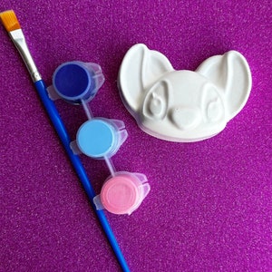 May include: A white ceramic figurine shaped like a cartoon dog with three paint pots: blue, light blue, and pink.