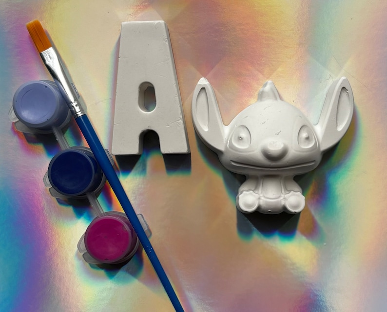 May include: A white ceramic letter A and a white ceramic figurine of Stitch from Lilo & Stitch, both unpainted. Three paint pots with lids are included: one light blue, one dark blue, and one magenta.