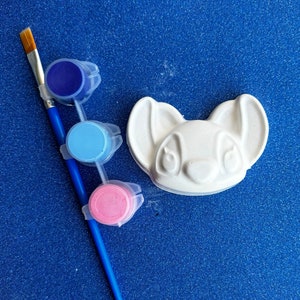 May include: A white ceramic figurine of a cartoon character with large ears and eyes. The figurine is accompanied by three small paint pots with blue, pink, and light blue paint, and a paintbrush.