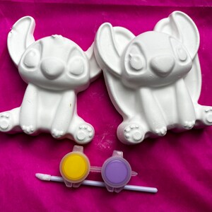 May include: Two white ceramic figurines of Stitch from Lilo & Stitch, ready to be painted. The figurines are sitting on a pink surface.  A yellow and purple paint pot with a brush is included.