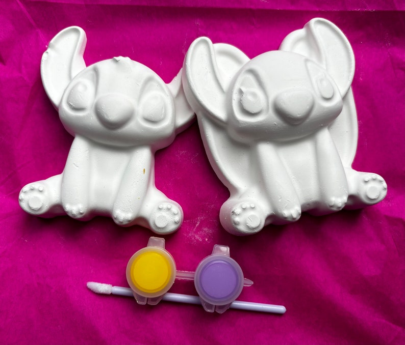 May include: Two white ceramic figurines of Stitch from Lilo & Stitch, ready to be painted. The figurines are sitting on a pink surface.  A yellow and purple paint pot with a brush is included.