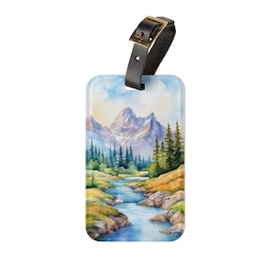 Op de afbeelding: Bagagelabel met een aquarel landschap van bergen, een rivier en groenblijvende bomen. Het heeft een zwarte leren riem met een goudkleurige gesp. De achtergrond is een lichtblauwe lucht met wolken.