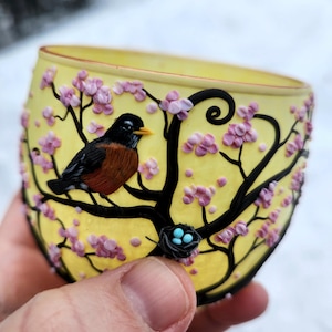 Puede incluir: Vaso amarillo con un dise&ntilde;o detallado de un p&aacute;jaro posado en una rama con flores rosas. El p&aacute;jaro tiene cabeza negra, pecho marr&oacute;n y pico naranja. Tambi&eacute;n se ve un peque&ntilde;o nido con huevos azules.