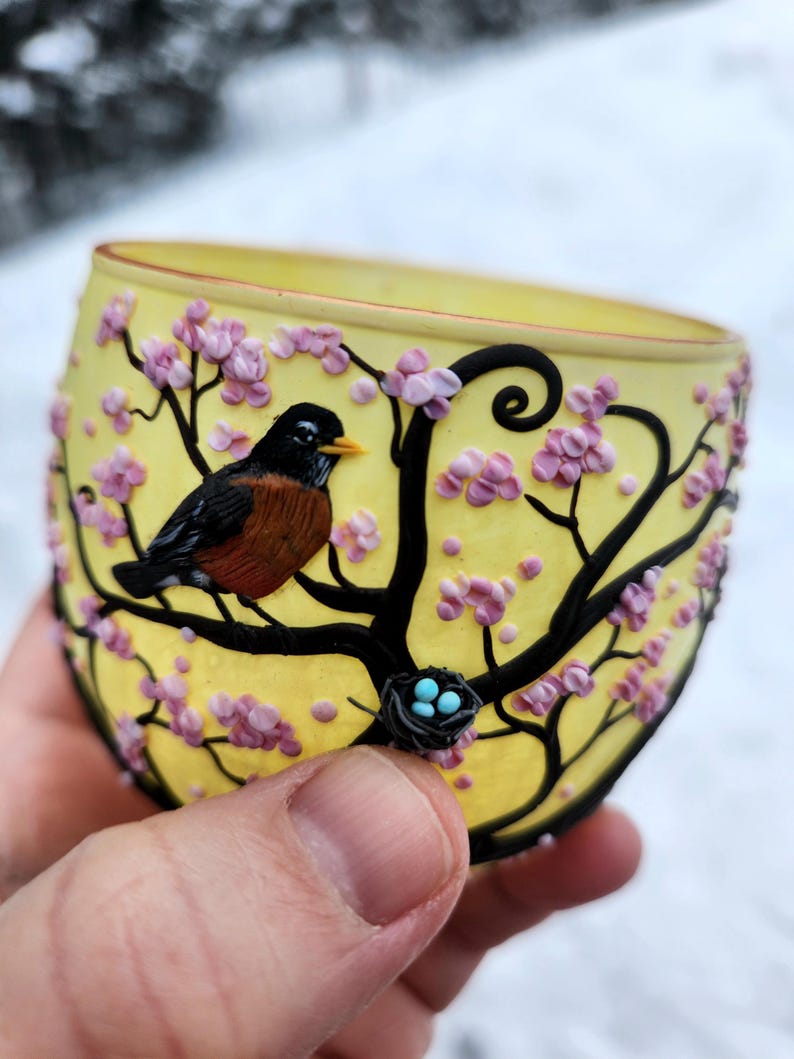 Puede incluir: Vaso amarillo con un dise&ntilde;o detallado de un p&aacute;jaro posado en una rama con flores rosas. El p&aacute;jaro tiene cabeza negra, pecho marr&oacute;n y pico naranja. Tambi&eacute;n se ve un peque&ntilde;o nido con huevos azules.