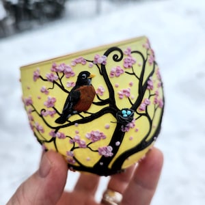 Puede incluir: Un vaso de vidrio amarillo con un dise&ntilde;o de &aacute;rbol negro, flores rosas y un petirrojo. El petirrojo est&aacute; posado en una rama cerca de un nido con tres huevos azules. El vaso se sostiene en una mano sobre un fondo nevado.