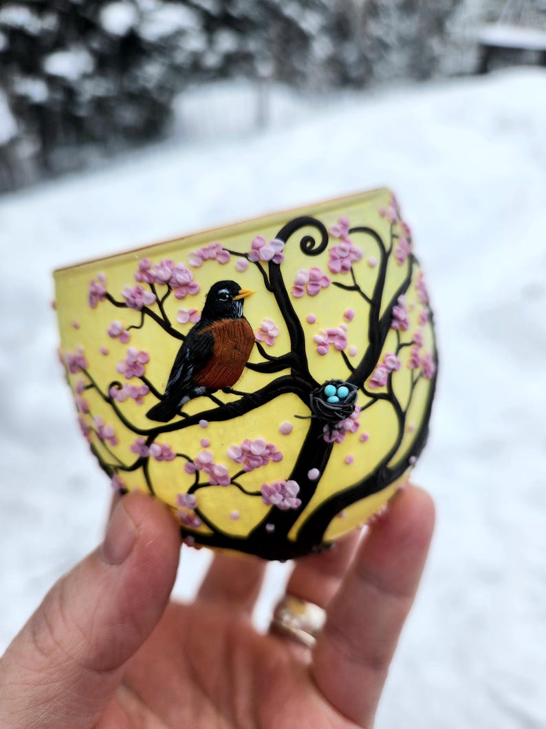Puede incluir: Un vaso de vidrio amarillo con un dise&ntilde;o de &aacute;rbol negro, flores rosas y un petirrojo. El petirrojo est&aacute; posado en una rama cerca de un nido con tres huevos azules. El vaso se sostiene en una mano sobre un fondo nevado.