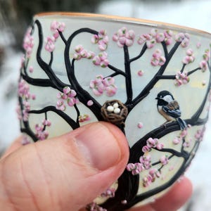 Puede incluir: Un vaso pintado a mano con un dise&ntilde;o tem&aacute;tico de la naturaleza. El vaso presenta un &aacute;rbol negro con flores rosas, un p&aacute;jaro posado en una rama y un peque&ntilde;o nido con huevos. El borde del vaso tiene un ribete dorado.