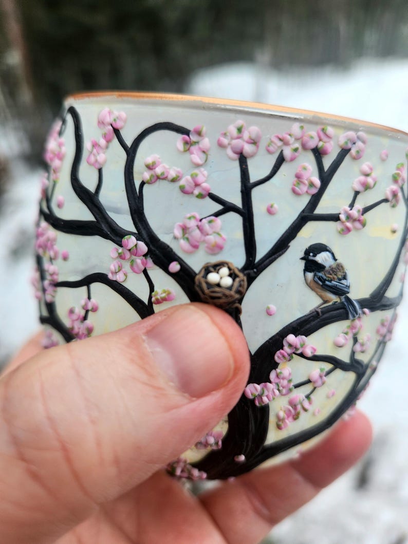 Puede incluir: Un vaso pintado a mano con un dise&ntilde;o tem&aacute;tico de la naturaleza. El vaso presenta un &aacute;rbol negro con flores rosas, un p&aacute;jaro posado en una rama y un peque&ntilde;o nido con huevos. El borde del vaso tiene un ribete dorado.