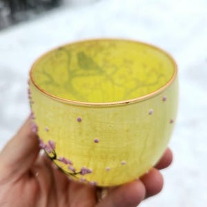 Puede incluir: Una taza de t&eacute; de vidrio amarillo con un dise&ntilde;o floral y un borde dorado. La taza est&aacute; decorada con peque&ntilde;os puntos rosas y una rama de flores rosas. La taza se sostiene en una mano.