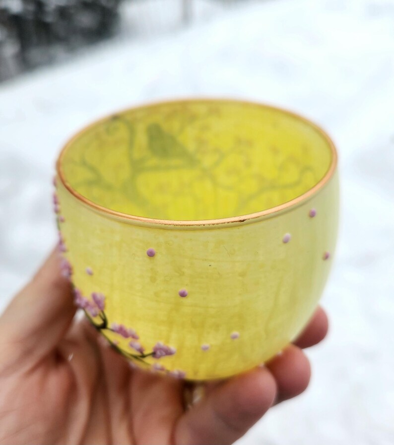 Puede incluir: Una taza de t&eacute; de vidrio amarillo con un dise&ntilde;o floral y un borde dorado. La taza est&aacute; decorada con peque&ntilde;os puntos rosas y una rama de flores rosas. La taza se sostiene en una mano.