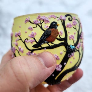 Puede incluir: Un portavelas de vidrio amarillo con un dise&ntilde;o detallado de un petirrojo posado en una rama con flores rosas. El p&aacute;jaro tiene el pecho marr&oacute;n y alas negras. Tambi&eacute;n es visible un peque&ntilde;o nido con huevos azules.