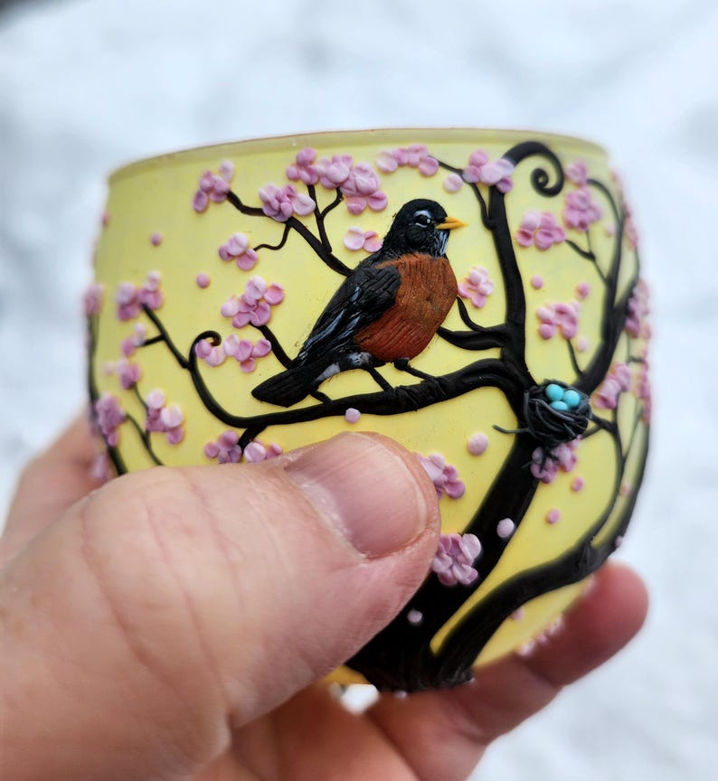 Puede incluir: Un portavelas de vidrio amarillo con un dise&ntilde;o detallado de un petirrojo posado en una rama con flores rosas. El p&aacute;jaro tiene el pecho marr&oacute;n y alas negras. Tambi&eacute;n es visible un peque&ntilde;o nido con huevos azules.