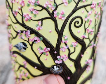 Una mamá carbonera y su nido con huevos en un árbol en flor de primavera, esculpidos con arcilla polimérica sobre un portavelas de vidrio reciclado en color amarillo.