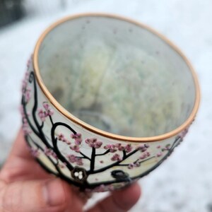 Puede incluir: Un cuenco de vidrio transparente con un borde dorado y un dise&ntilde;o floral pintado. El cuenco presenta ramas negras con flores rosas. Un peque&ntilde;o p&aacute;jaro est&aacute; posado en una rama. El cuenco se sostiene en una mano.