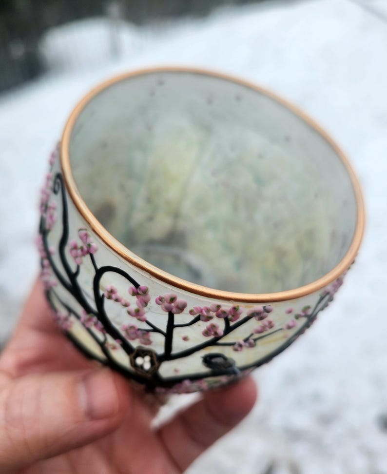 Puede incluir: Un cuenco de vidrio transparente con un borde dorado y un dise&ntilde;o floral pintado. El cuenco presenta ramas negras con flores rosas. Un peque&ntilde;o p&aacute;jaro est&aacute; posado en una rama. El cuenco se sostiene en una mano.