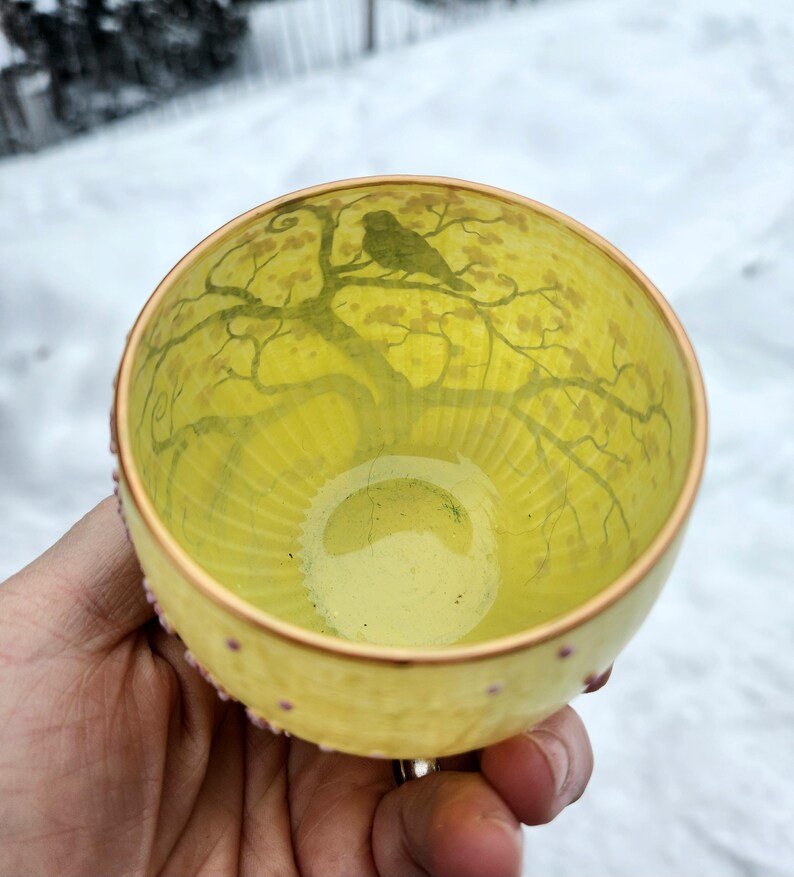 Puede incluir: Un cuenco de vidrio amarillo con una escena interior pintada de un &aacute;rbol y un p&aacute;jaro. El borde es dorado con peque&ntilde;os puntos rosas. El cuenco se sostiene en una mano sobre un fondo nevado.