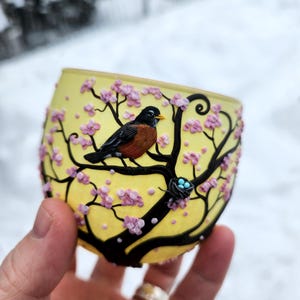 Puede incluir: Un portavelas de cristal amarillo con un dise&ntilde;o de &aacute;rbol negro, flores rosas y un petirrojo. Tambi&eacute;n se ve un peque&ntilde;o nido con tres huevos azules. El portavelas se sostiene en una mano.