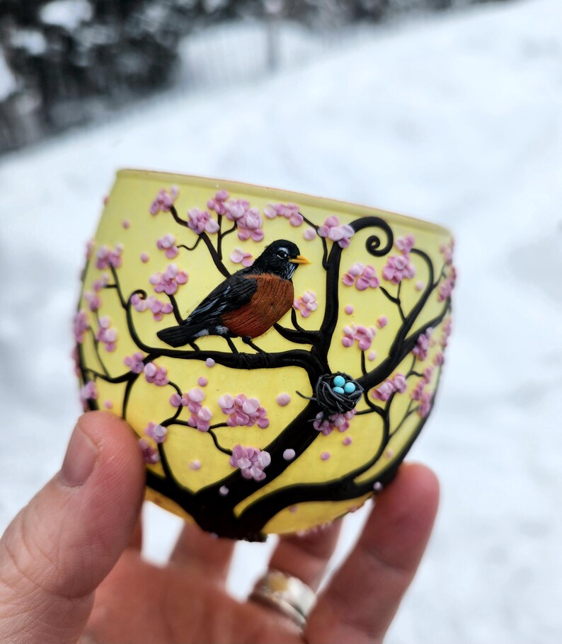 Puede incluir: Un portavelas de cristal amarillo con un dise&ntilde;o de &aacute;rbol negro, flores rosas y un petirrojo. Tambi&eacute;n se ve un peque&ntilde;o nido con tres huevos azules. El portavelas se sostiene en una mano.