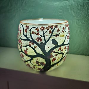 Puede incluir: Un portavelas de cristal con un dise&ntilde;o de &aacute;rbol. El &aacute;rbol tiene ramas negras, bayas rojas y un peque&ntilde;o p&aacute;jaro posado en una rama. El portavelas est&aacute; iluminado desde dentro, creando un brillo c&aacute;lido.