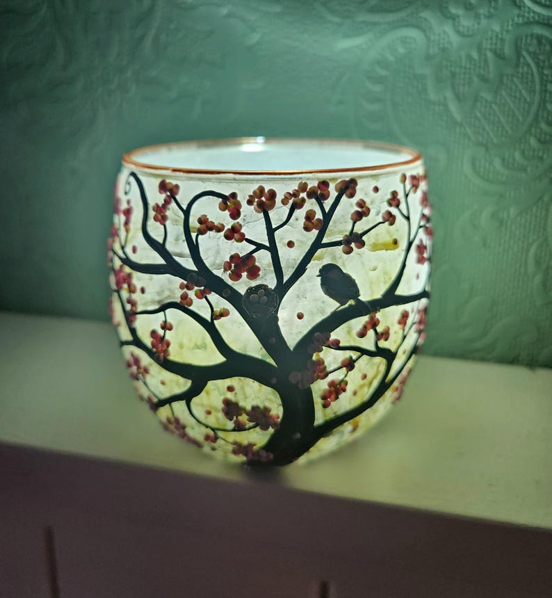 Puede incluir: Un portavelas de cristal con un dise&ntilde;o de &aacute;rbol. El &aacute;rbol tiene ramas negras, bayas rojas y un peque&ntilde;o p&aacute;jaro posado en una rama. El portavelas est&aacute; iluminado desde dentro, creando un brillo c&aacute;lido.