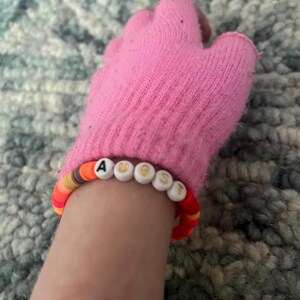 May include: A pink knit fingerless glove with a beaded bracelet that spells "AUGUST" in white letters with gold trim.