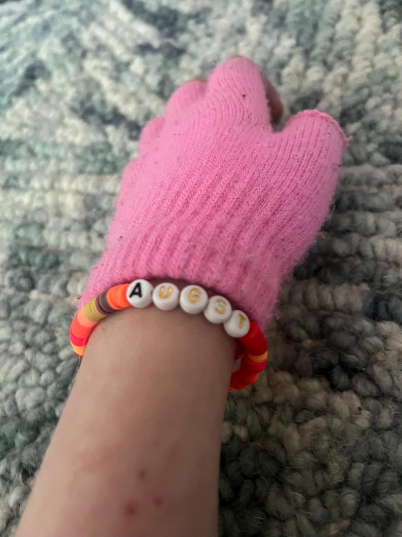 May include: A pink knit fingerless glove with a beaded bracelet that spells "AUGUST" in white letters with gold trim.
