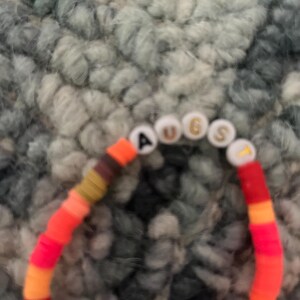 May include: A multi-colored beaded bracelet with the word "AUGUST" spelled out in white letters on white beads.