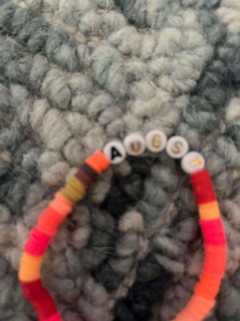 May include: A multi-colored beaded bracelet with the word "AUGUST" spelled out in white letters on white beads.