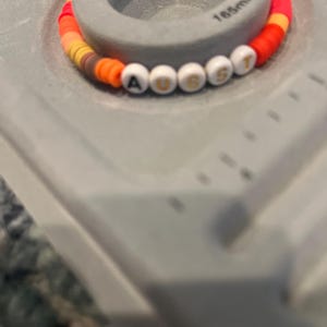 May include: A colorful beaded bracelet with white letter beads spelling out "AUGUST". The bracelet is made with orange, red, yellow, and brown beads.
