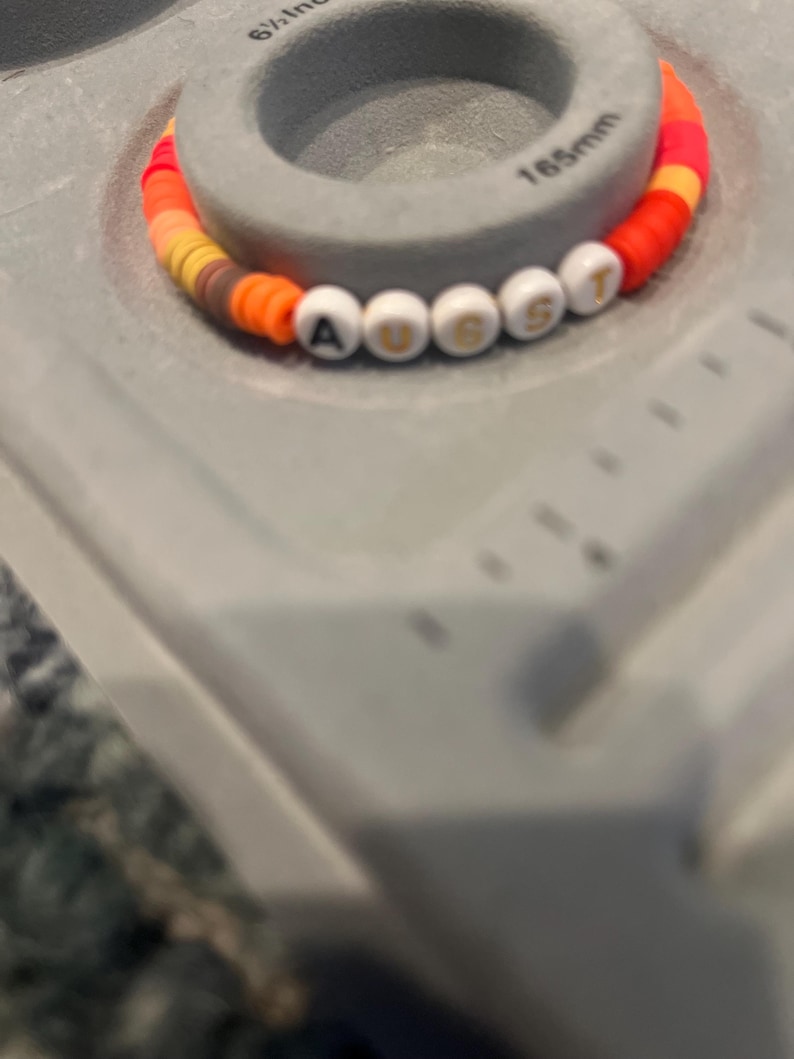May include: A colorful beaded bracelet with white letter beads spelling out "AUGUST". The bracelet is made with orange, red, yellow, and brown beads.