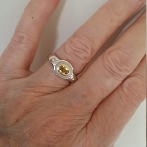K&ouml;nnte beinhalten: Ein silberner Ring mit einem gelben ovalen Edelstein, der in einer Zargenfassung gefasst ist. Der Ring hat zwei kleine silberne Kugeln auf jeder Seite des Edelsteins.