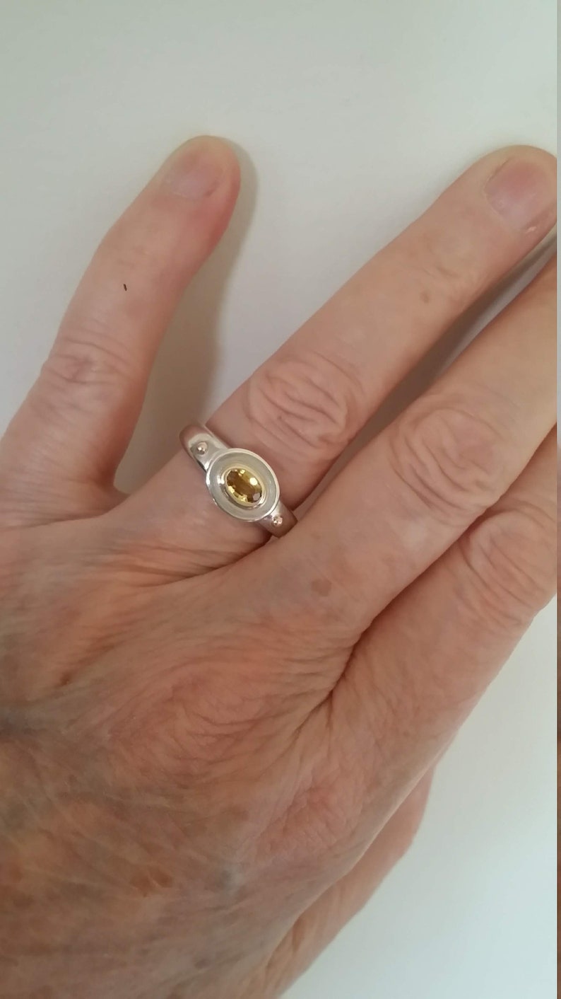 K&ouml;nnte beinhalten: Ein silberner Ring mit einem gelben ovalen Edelstein, der in einer Zargenfassung gefasst ist. Der Ring hat zwei kleine silberne Kugeln auf jeder Seite des Edelsteins.