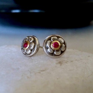 May include: A pair of silver stud earrings with a red gemstone in the centre of each earring. The earrings are surrounded by a gold rim and have a floral design.