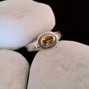 K&ouml;nnte beinhalten: Ein silberner Ring mit einer goldenen Fassung und einem gelben ovalen Edelstein. Der Ring liegt auf wei&szlig;en Steinen.