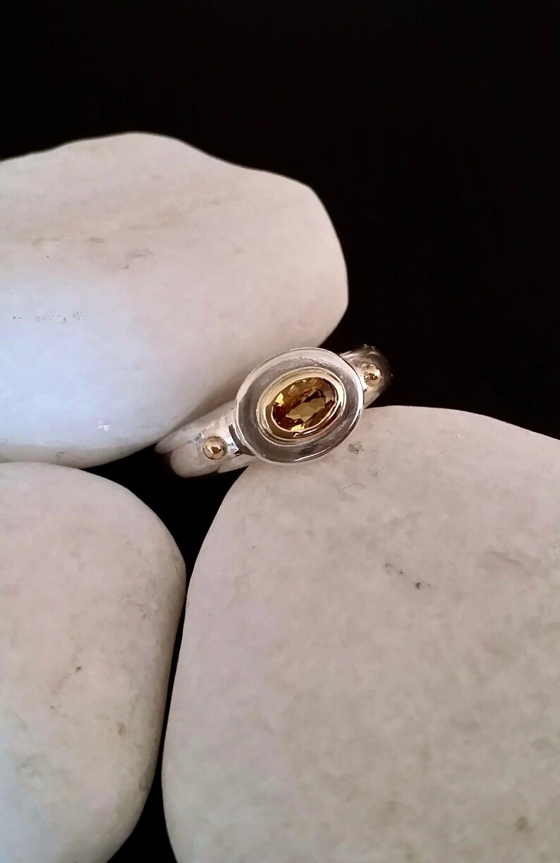 K&ouml;nnte beinhalten: Ein silberner Ring mit einer goldenen Fassung und einem gelben ovalen Edelstein. Der Ring liegt auf wei&szlig;en Steinen.