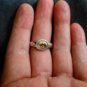 K&ouml;nnte beinhalten: Ein silberner Ring mit einem gelben ovalen Edelstein, der in einer goldenen Fassung sitzt. Der Ring hat zwei kleine silberne Kugeln auf jeder Seite des Edelsteins.