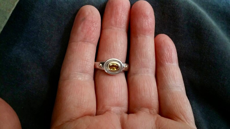 K&ouml;nnte beinhalten: Ein silberner Ring mit einem gelben ovalen Edelstein, der in einer goldenen Fassung sitzt. Der Ring hat zwei kleine silberne Kugeln auf jeder Seite des Edelsteins.