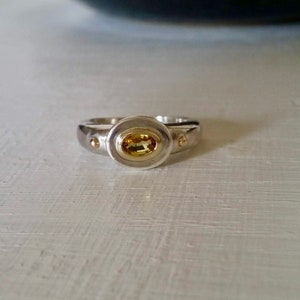 K&ouml;nnte beinhalten: Ein silberner Ring mit einem gelben ovalen Edelstein, der in einer goldenen Fassung sitzt. Der Ring hat zwei kleine goldene Akzente auf beiden Seiten des Edelsteins.