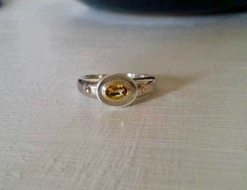 K&ouml;nnte beinhalten: Ein silberner Ring mit einem gelben ovalen Edelstein, der in einer goldenen Fassung sitzt. Der Ring hat zwei kleine goldene Akzente auf beiden Seiten des Edelsteins.