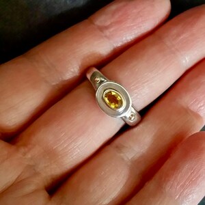 K&ouml;nnte beinhalten: Ein silberner Ring mit einem gelben ovalen Edelstein, der in einer goldenen Fassung sitzt. Der Ring hat zwei kleine goldene Punkte auf der Ringschiene.