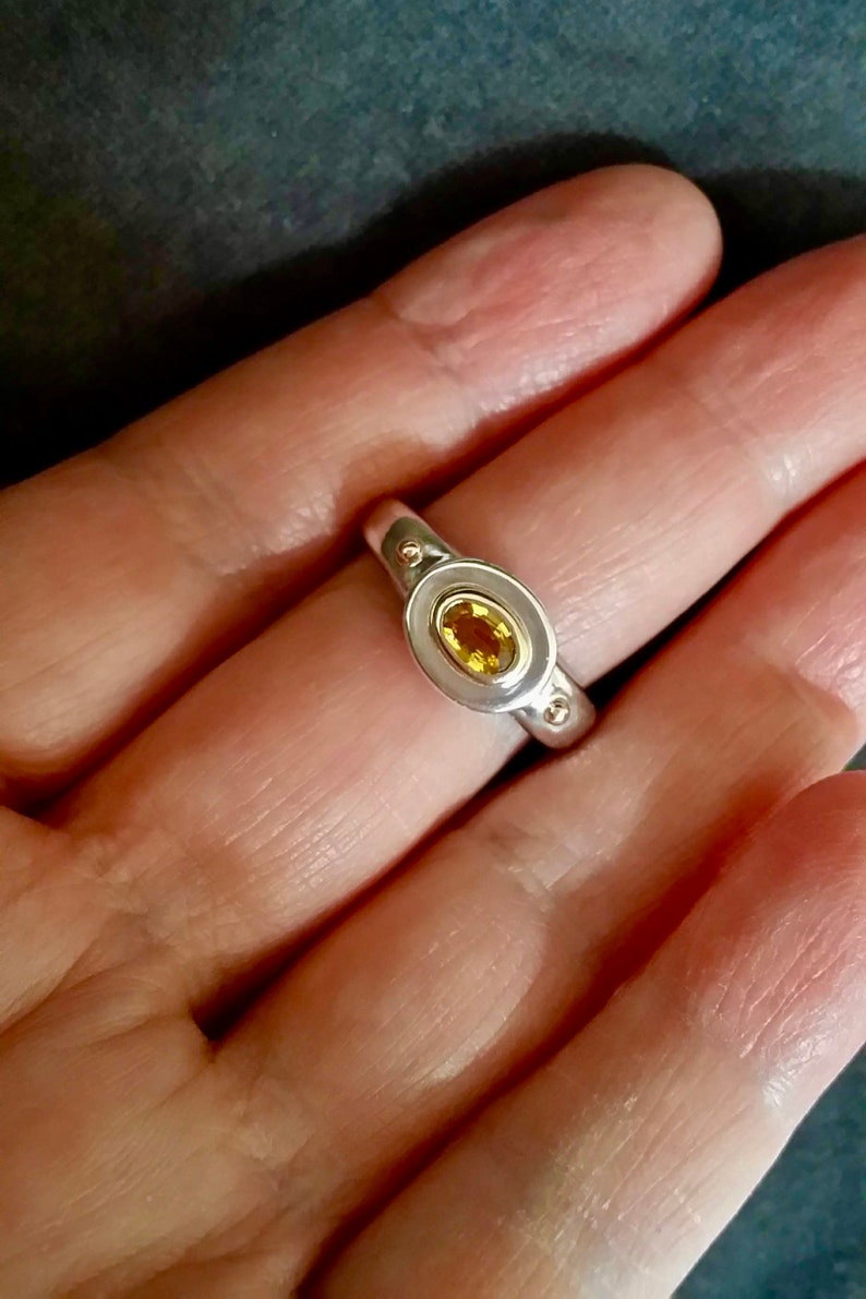 K&ouml;nnte beinhalten: Ein silberner Ring mit einem gelben ovalen Edelstein, der in einer goldenen Fassung sitzt. Der Ring hat zwei kleine goldene Punkte auf der Ringschiene.