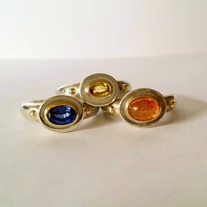 K&ouml;nnte beinhalten: Drei Silberringe mit goldenen Akzenten und ovalen Edelsteinen. Die Ringe zeigen einen blauen Saphir, einen gelben Saphir und einen orangefarbenen Sonnen-Stein.