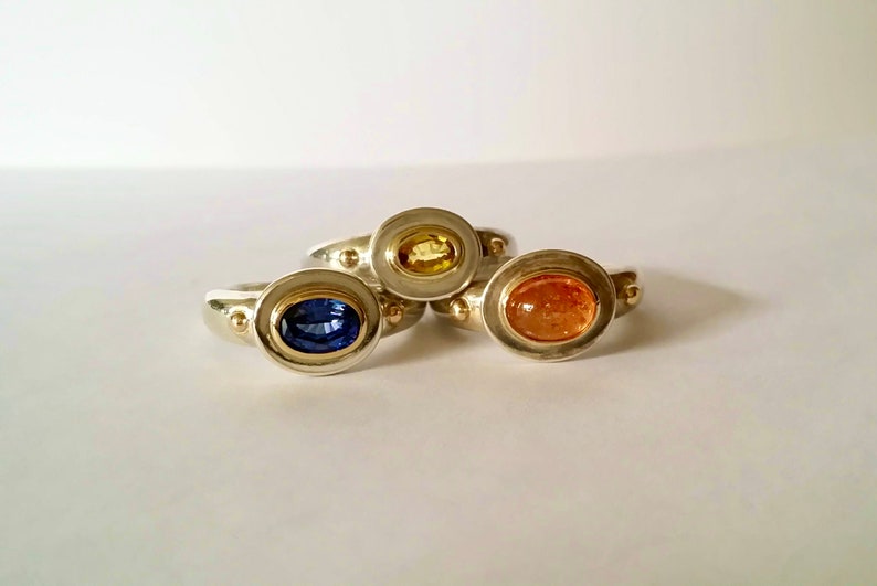 K&ouml;nnte beinhalten: Drei Silberringe mit goldenen Akzenten und ovalen Edelsteinen. Die Ringe zeigen einen blauen Saphir, einen gelben Saphir und einen orangefarbenen Sonnen-Stein.