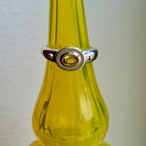 K&ouml;nnte beinhalten: Ein silberner Ring mit einem gelben ovalen Edelstein, der in einer Zargenfassung gefasst ist. Der Ring befindet sich auf einer gelben Glasflasche.