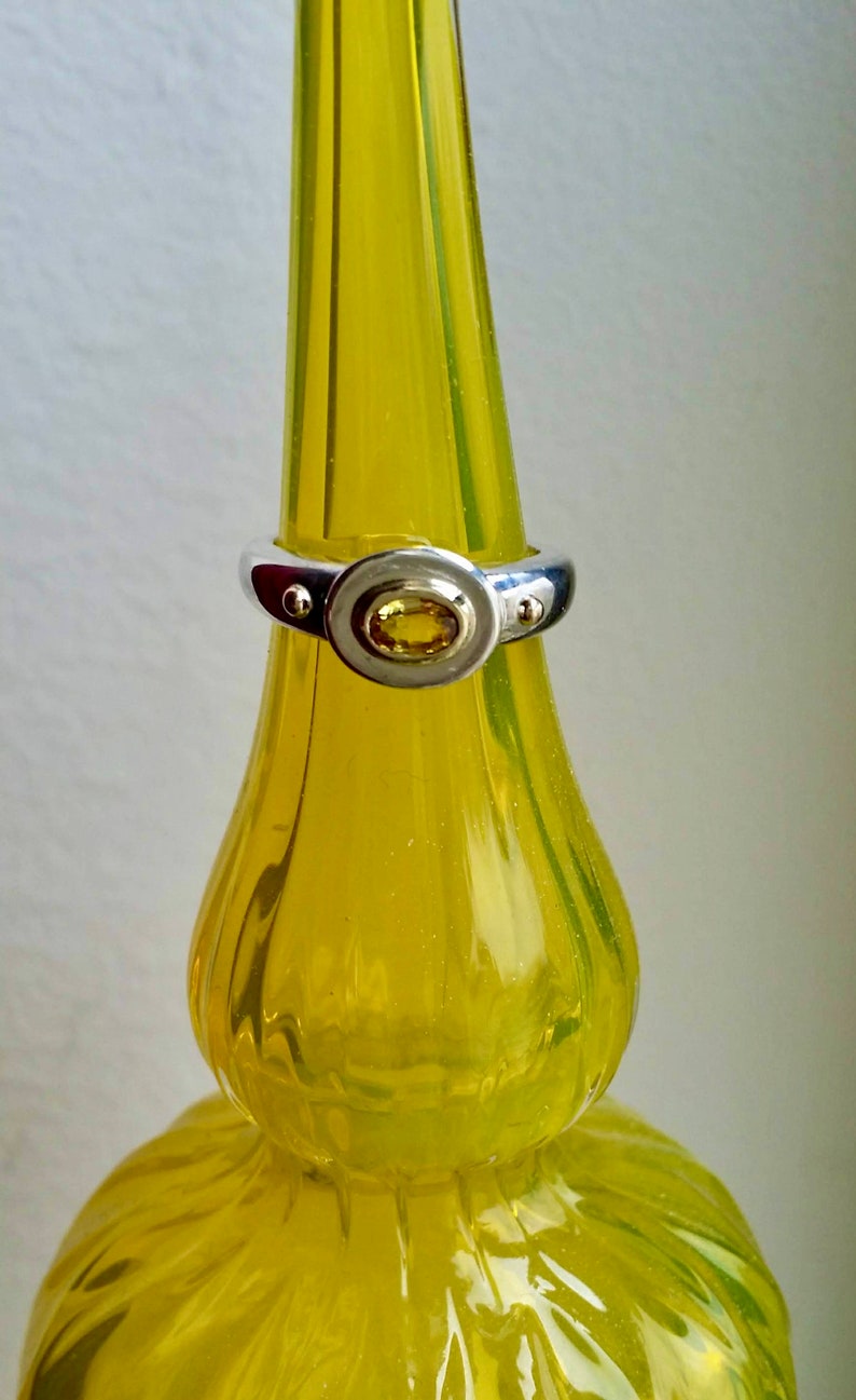 K&ouml;nnte beinhalten: Ein silberner Ring mit einem gelben ovalen Edelstein, der in einer Zargenfassung gefasst ist. Der Ring befindet sich auf einer gelben Glasflasche.