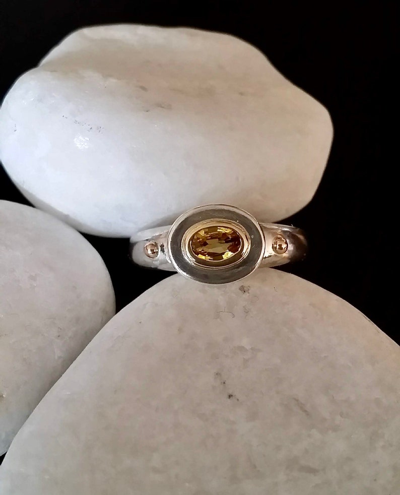 K&ouml;nnte beinhalten: Ein silberner Ring mit einem gelben ovalen Edelstein, der in einer Zargenfassung gefasst ist. Der Ring hat zwei kleine goldene Punkte auf jeder Seite des Steins.
