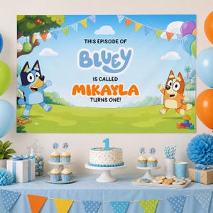 Op de afbeelding: Een Bluey-thema verjaardagsfeestje. Een banner leest "This episode of Bluey is called Mikayla turns one!" met Bluey en Bingo karakters. Een taart, cupcakes en lekkernijen op een blauwe tafel met ballonnen.